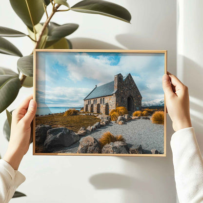 Hands holding framed Church of the Good Shepherd Lake Tekapo photography print in natural oak frame against white wall with large leaf plant