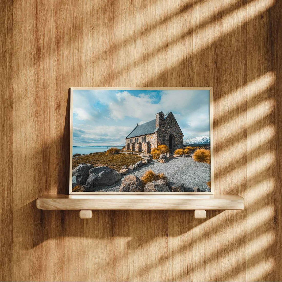 Framed Church of the Good Shepherd Lake Tekapo print in natural wood frame displayed on wooden shelf against warm oak panelled wall with dappled light