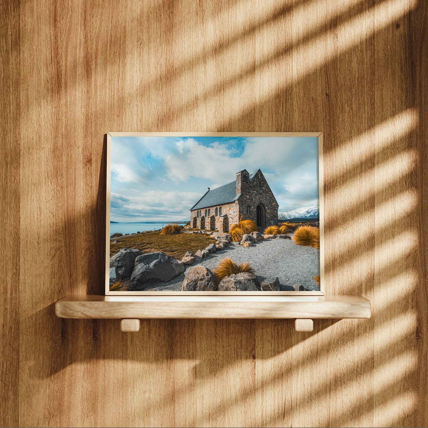 Framed Church of the Good Shepherd Lake Tekapo print in natural wood frame displayed on wooden shelf against warm oak panelled wall with dappled light