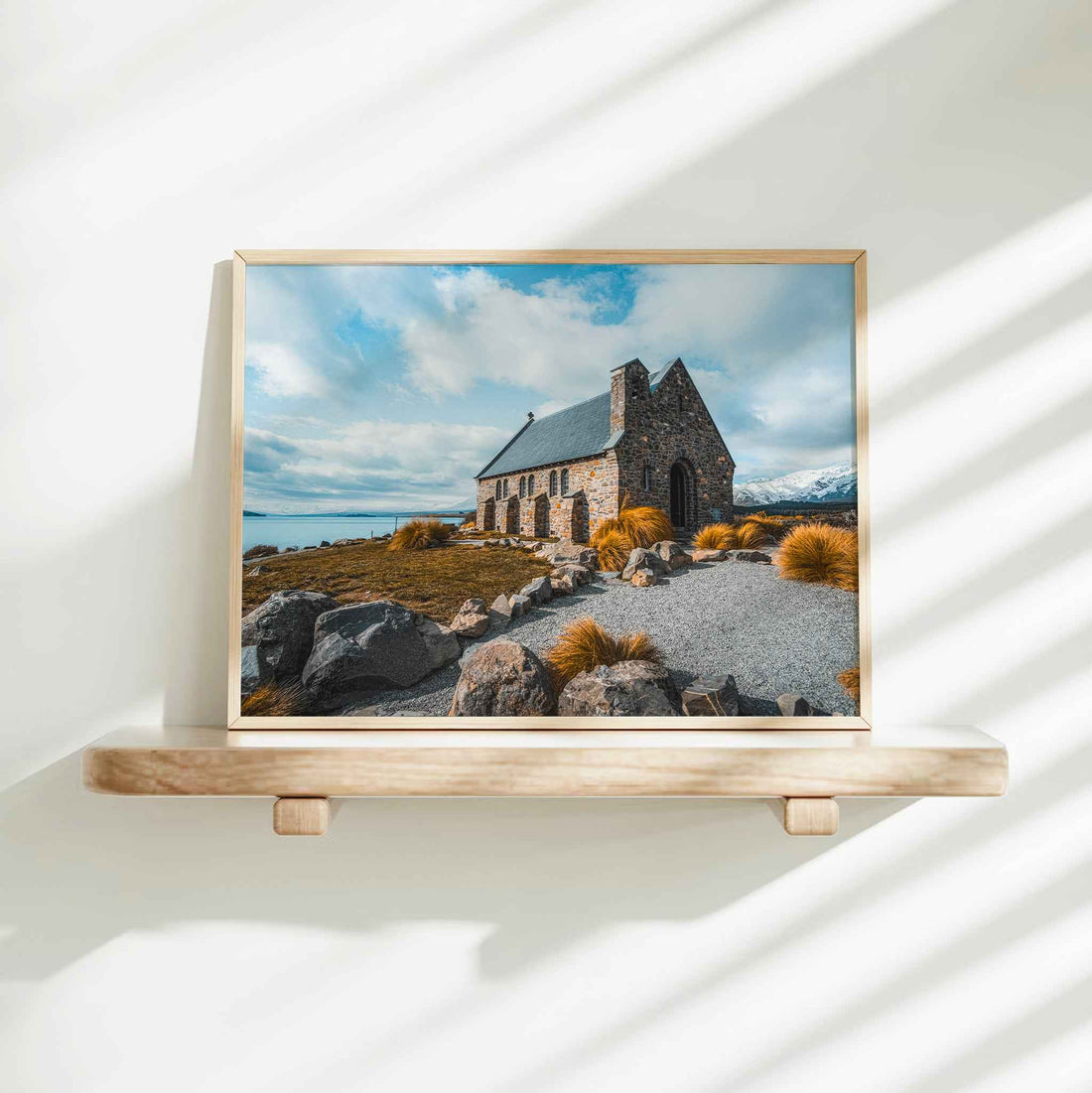 Framed Church of the Good Shepherd Lake Tekapo photography print in natural wood frame on minimalist wooden shelf against white wall with dappled shadows