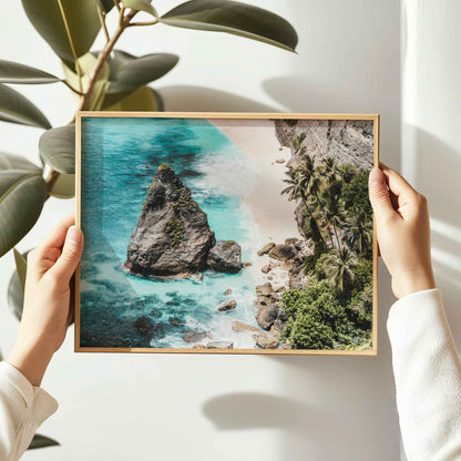 Hands holding Diamond Beach Nusa Penida landscape framed wall art print in natural oak wood frame against white wall with plant