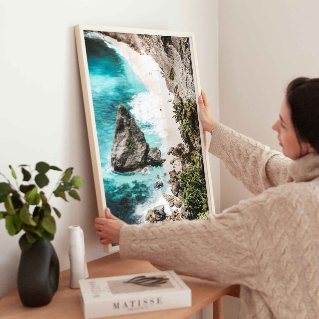 Woman holding large Diamond Beach Nusa Penida aerial framed print in natural wood frame above wooden side table with Matisse book and plant