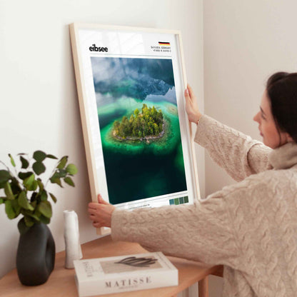 Woman placing large framed Eibsee aerial island Bavaria Germany print in natural wood frame on timber sideboard beside plant and books