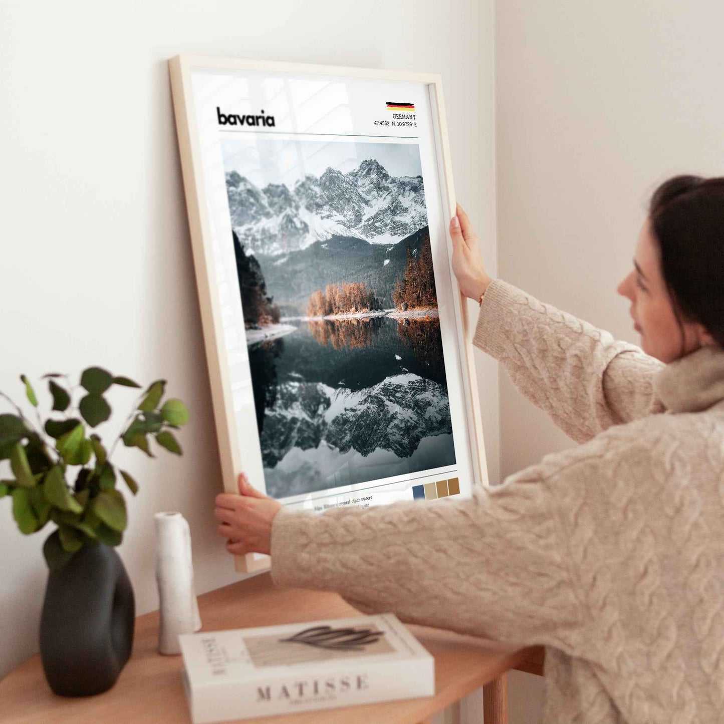 Woman placing large framed Eibsee Zugspitze Bavaria Germany print in natural wood frame on timber sideboard beside plant and books