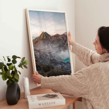 Woman holding large Ijen volcano crater lake Java Indonesia framed print in natural wood frame above wooden side table with Matisse book and plant