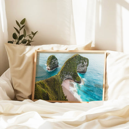 Natural wood framed Kelingking Beach Nusa Penida Bali aerial print leaning against pillows in a bedroom setting