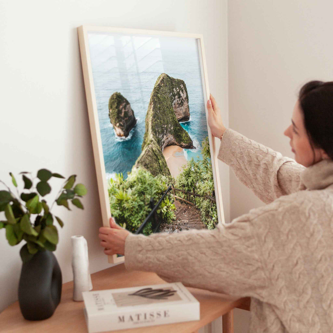 Woman holding large Kelingking Beach Nusa Penida framed print in natural wood frame above wooden side table with Matisse book and plant