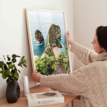 Woman holding large Kelingking Beach Nusa Penida framed print in natural wood frame above wooden side table with Matisse book and plant