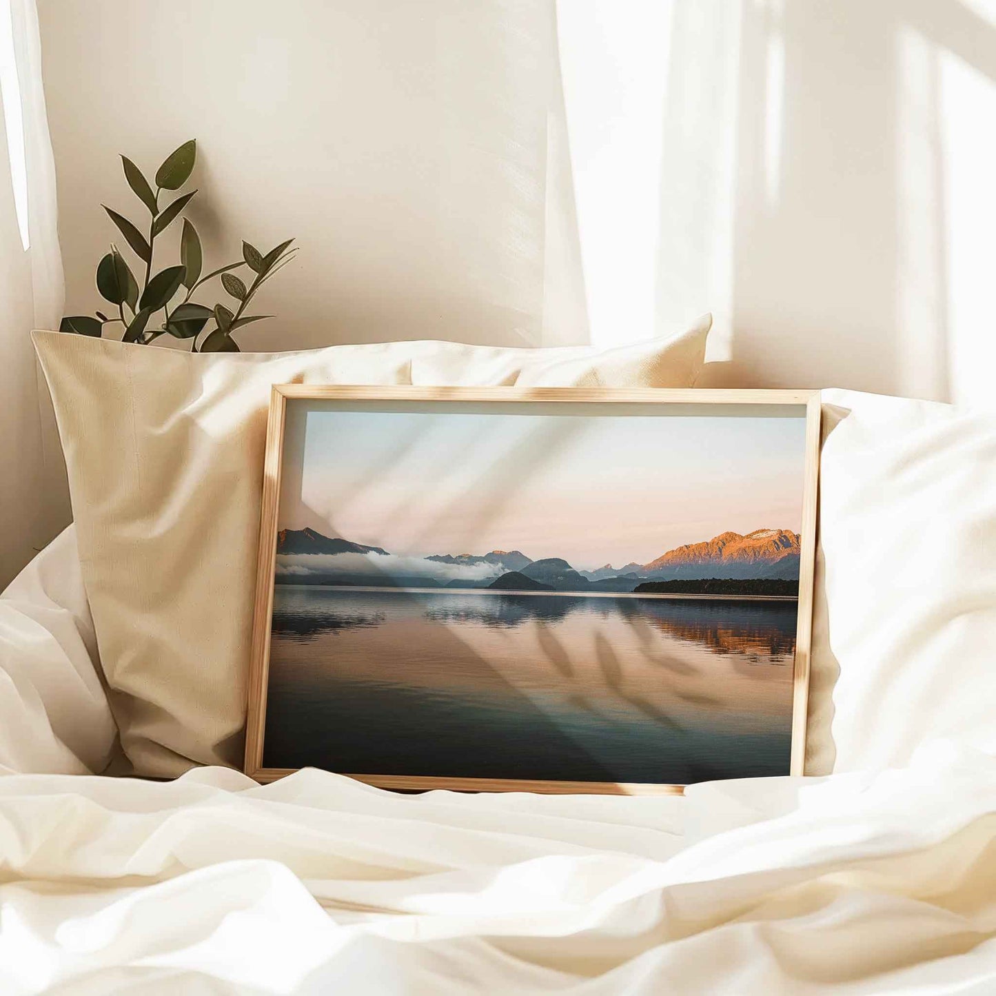 Natural wood framed Lake Manapouri New Zealand print leaning against cream pillow on white linen bed in soft morning light