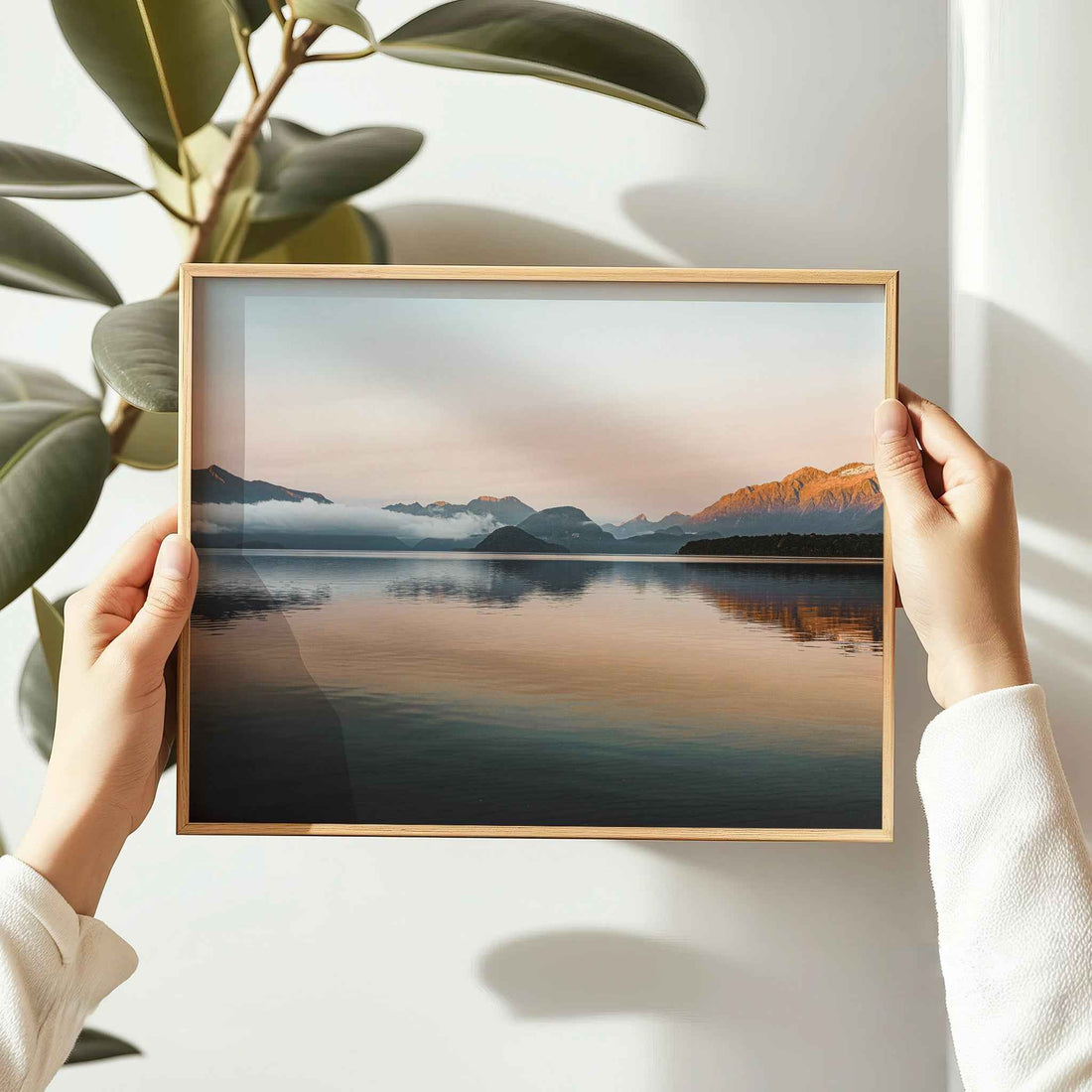 Hands holding framed Lake Manapouri New Zealand photography print in natural oak frame beside indoor plant