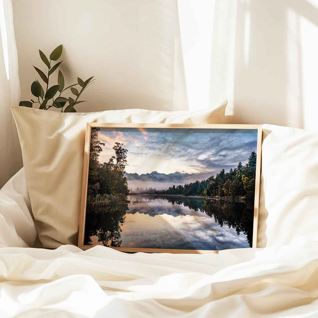 Framed Lake Matheson reflection sunrise print in natural wood frame resting against linen pillows on white bed with plant and curtains