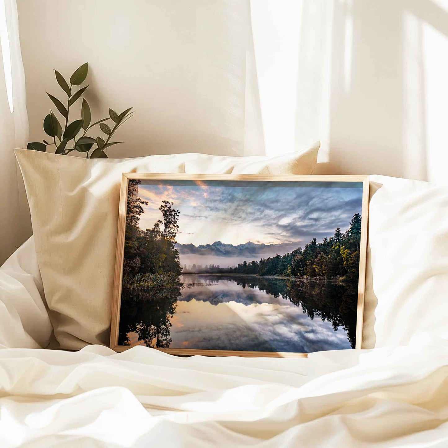 Framed Lake Matheson reflection sunrise print in natural wood frame resting against linen pillows on white bed with plant and curtains