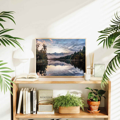Natural wood framed Lake Matheson reflection sunrise print displayed on wooden bookshelf in bright interior styled with plants and books