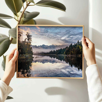 Hands holding framed Lake Matheson reflection sunrise photography print in natural oak frame against white wall with large leaf plant