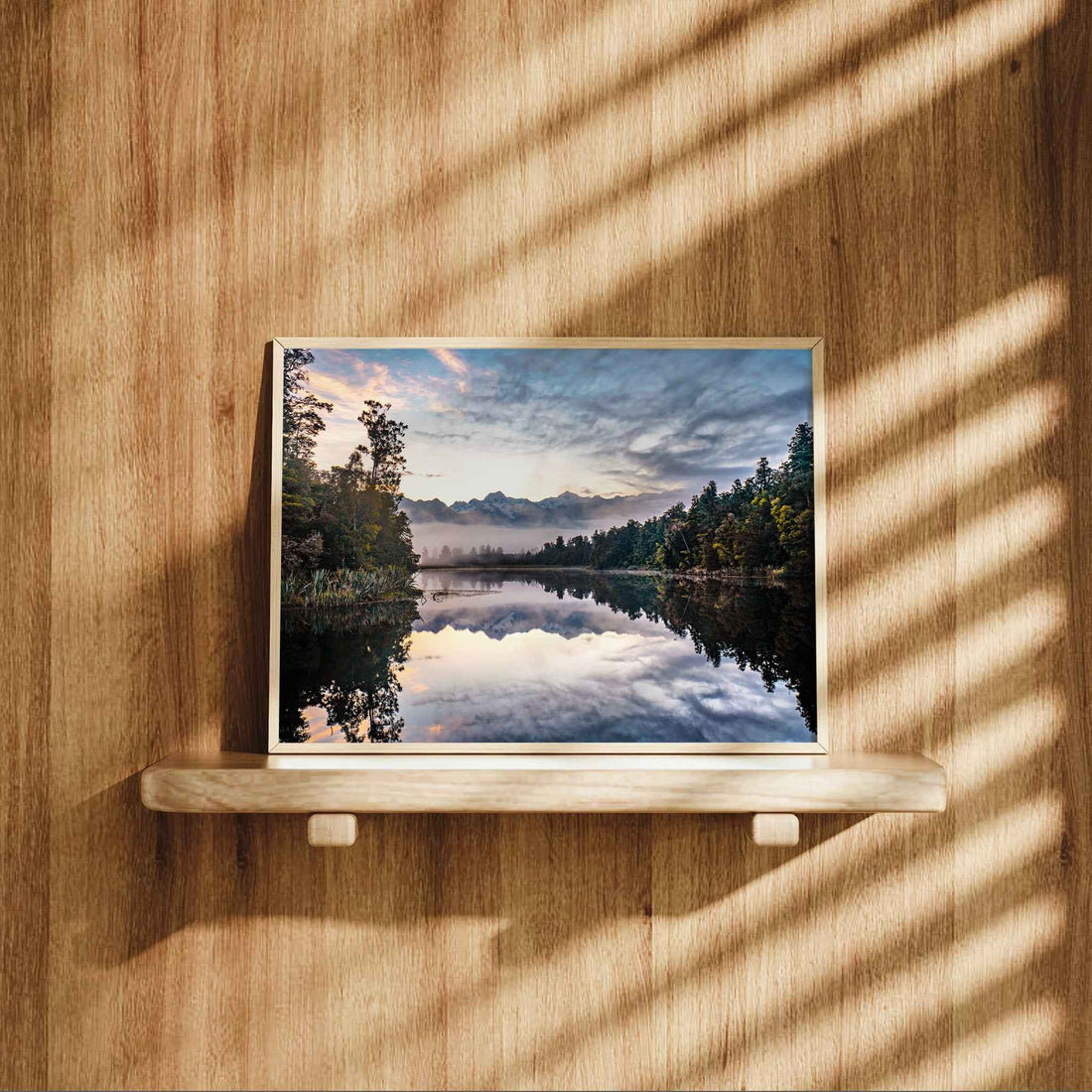 Framed Lake Matheson reflection sunrise print in natural wood frame displayed on wooden shelf against warm oak panelled wall with dappled light