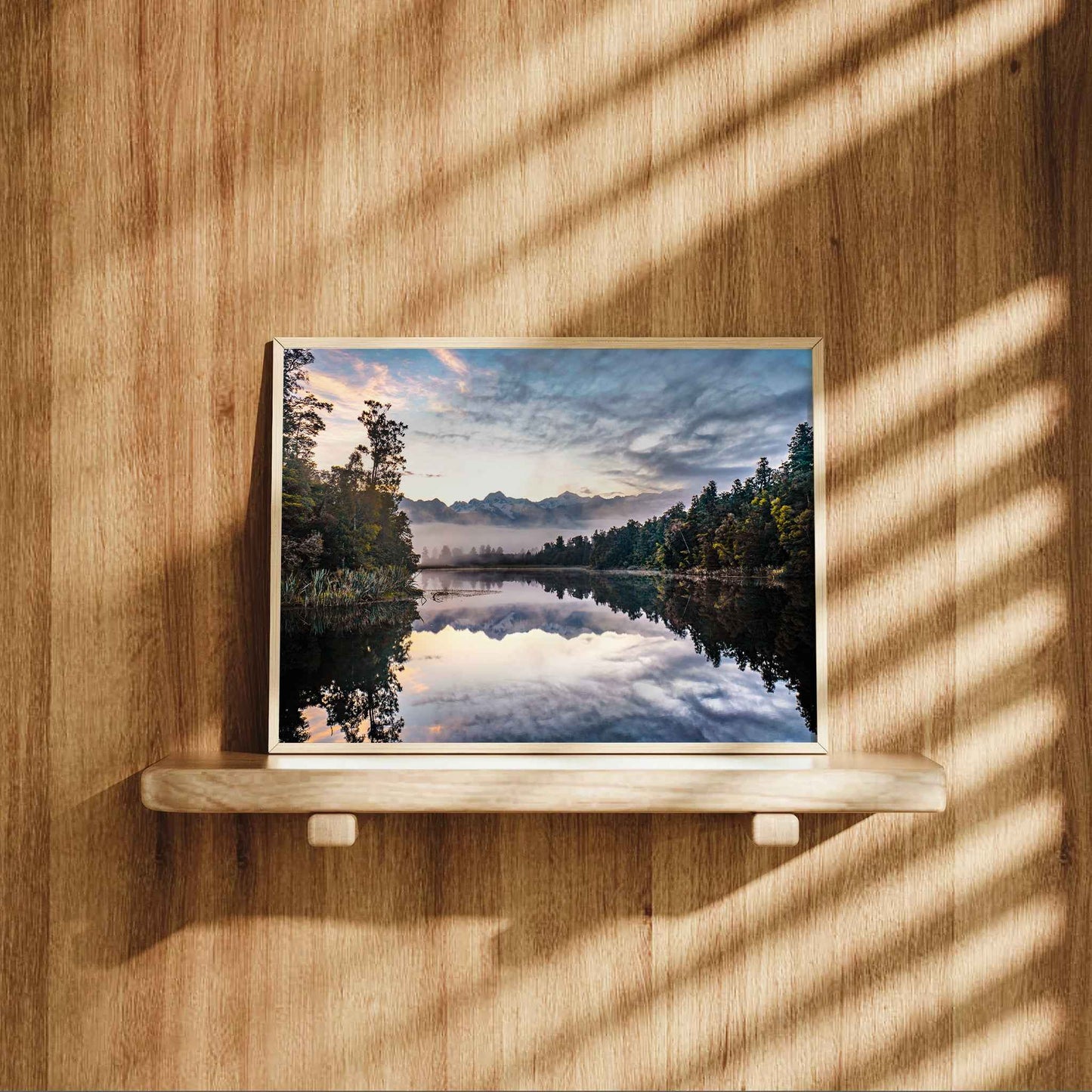 Framed Lake Matheson reflection sunrise print in natural wood frame displayed on wooden shelf against warm oak panelled wall with dappled light