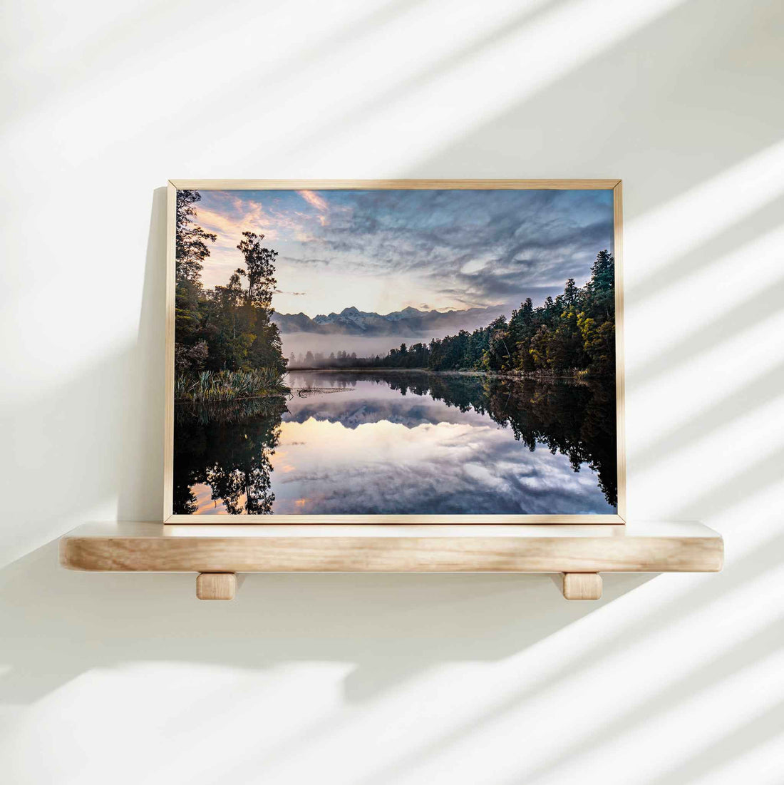 Framed Lake Matheson reflection sunrise photography print in natural wood frame on minimalist wooden shelf against white wall with dappled shadows