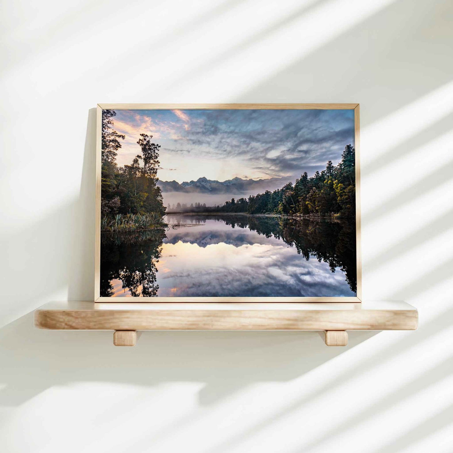 Framed Lake Matheson reflection sunrise photography print in natural wood frame on minimalist wooden shelf against white wall with dappled shadows