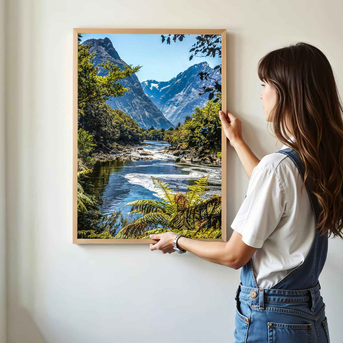 Woman hanging a natural wood framed Milford Track Fiordland New Zealand print on a white wall