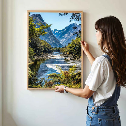 Woman hanging a natural wood framed Milford Track Fiordland New Zealand print on a white wall