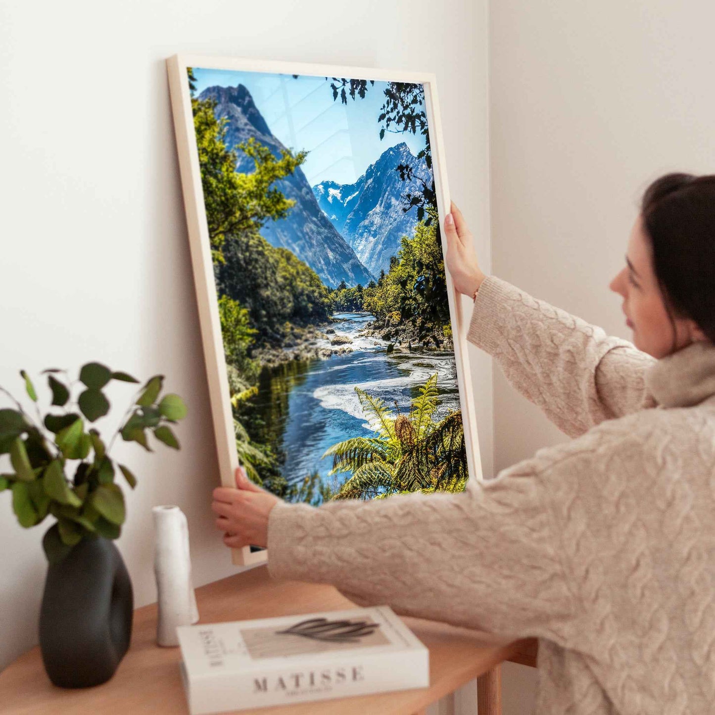Woman placing a natural wood framed Milford Track Fiordland New Zealand print on a wooden sideboard
