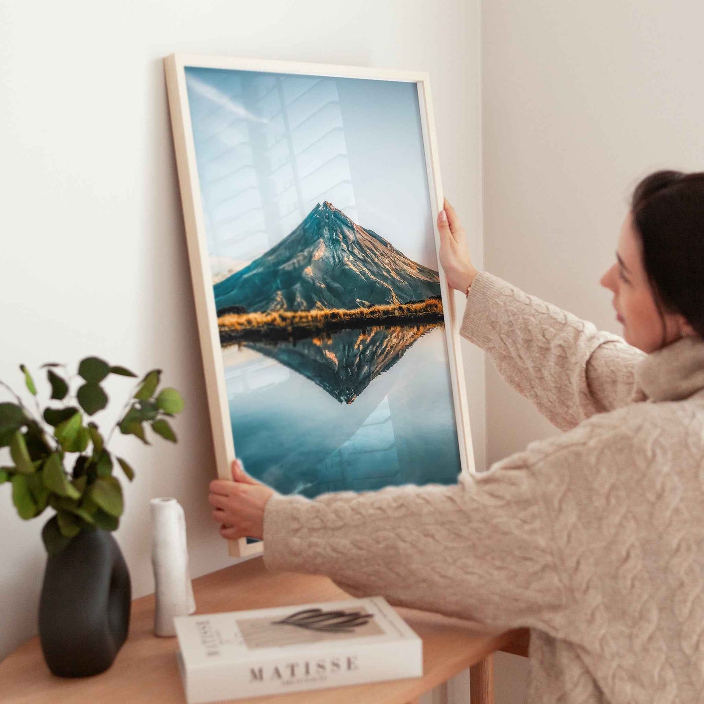 Woman holding large framed Mount Taranaki reflection photography print in natural wood frame near wooden table with plant and books