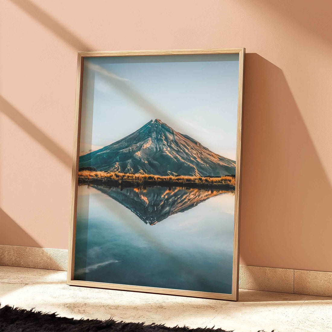 Framed Mount Taranaki lake reflection print in light wood frame leaning against terracotta wall with dramatic shadows