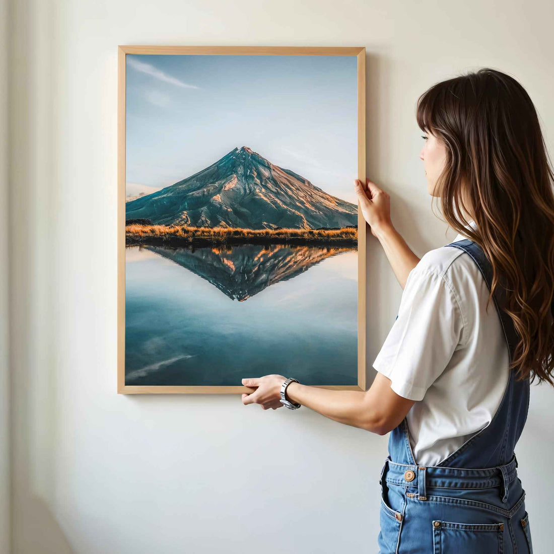 Woman hanging framed Mount Taranaki reflection photography print in natural oak frame on white wall