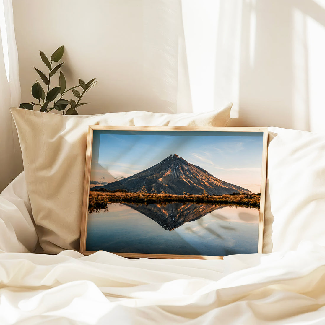 Natural wood framed Mount Taranaki reflection tarn print leaning against cream pillow on white linen bed in soft morning light