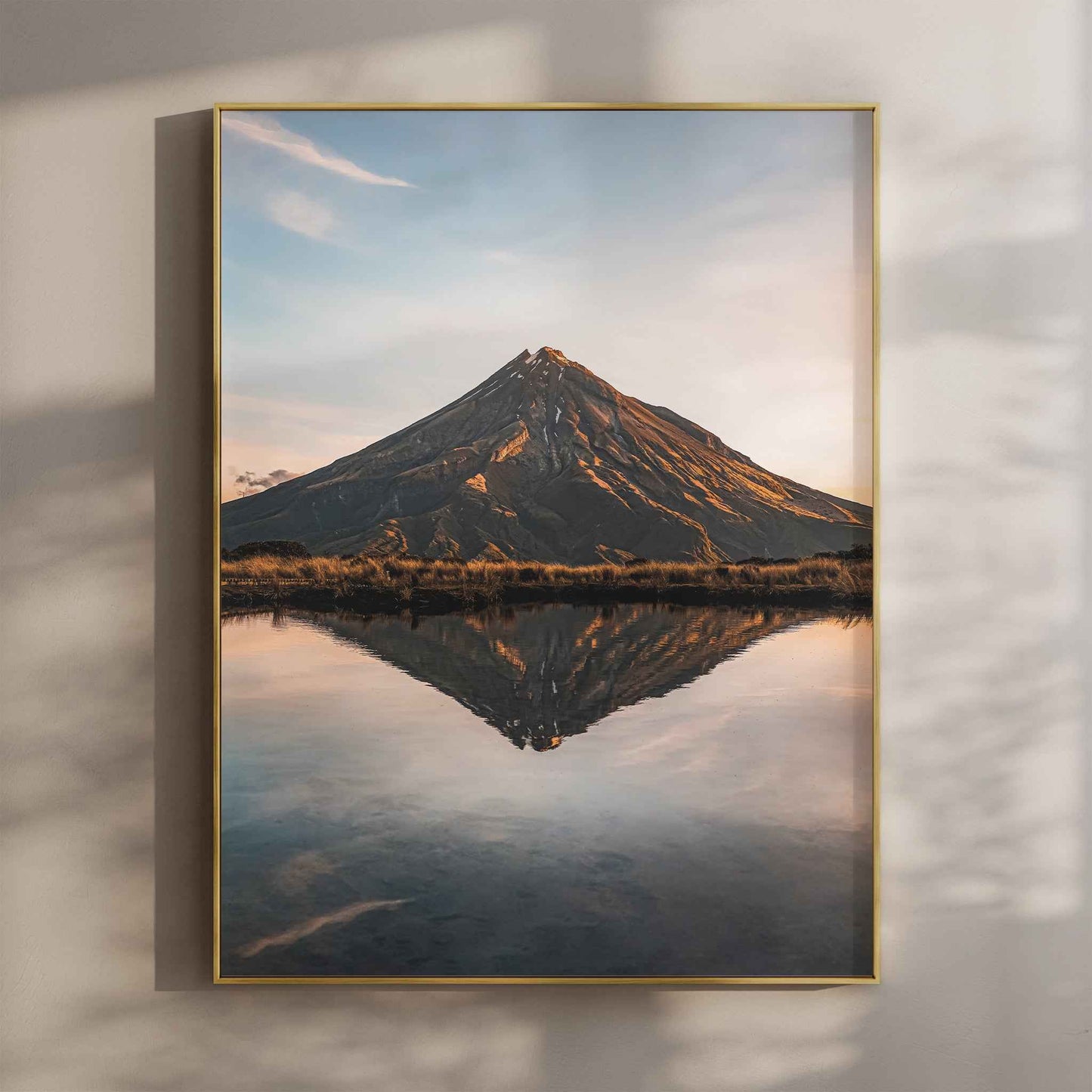 Framed Mount Taranaki sunset reflection photography print in gold float frame mounted on warm beige wall with window light shadows