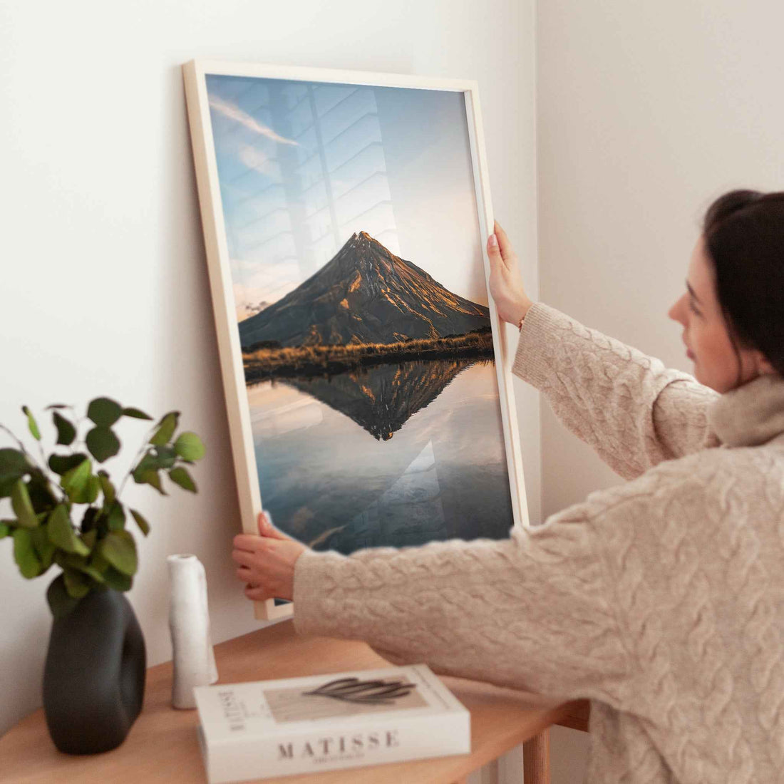 Woman holding large framed Mount Taranaki sunset reflection photography print in natural wood frame near wooden table with plant and books