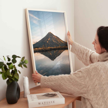Woman holding large framed Mount Taranaki sunset reflection photography print in natural wood frame near wooden table with plant and books
