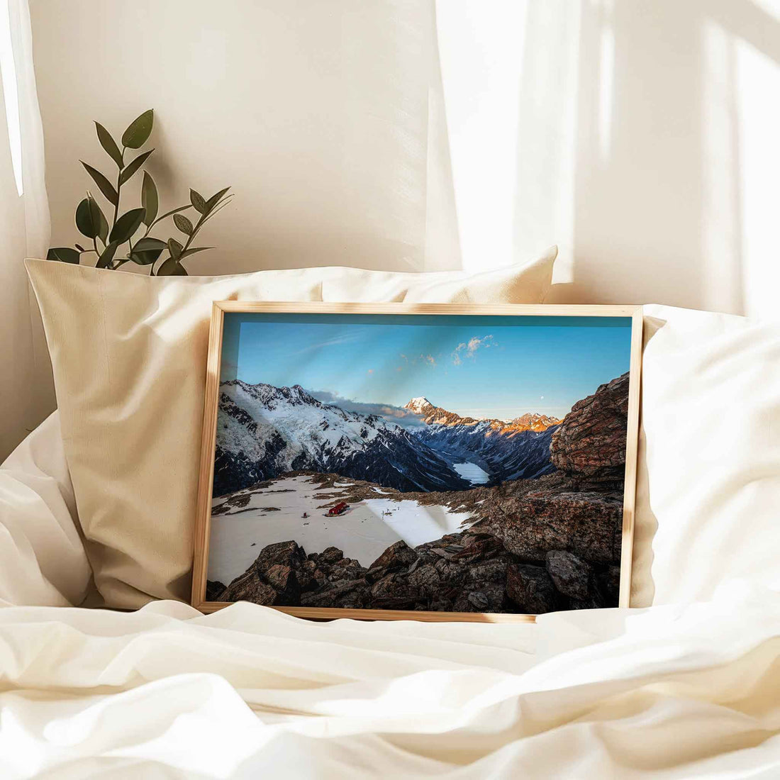 Natural wood framed Mueller Hut Mount Cook print leaning against cream pillow on white linen bed in soft morning light