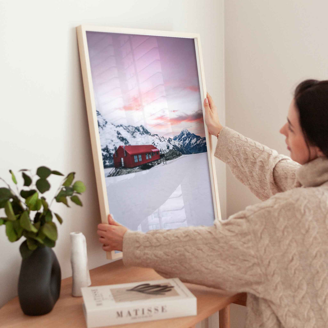 Woman holding large framed Mueller Hut and Mount Cook winter photography print in natural wood frame near wooden table with plant and books