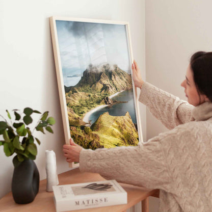 Woman holding large Padar Island Komodo National Park framed print in natural wood frame above wooden side table with Matisse book and plant