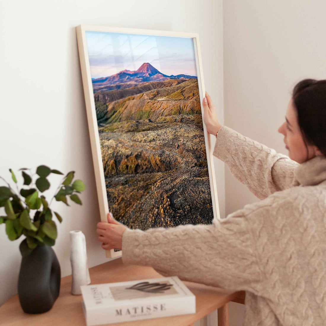 Woman holding large framed Mount Ruapehu landscape photography print in natural wood frame near wooden table with plant and books